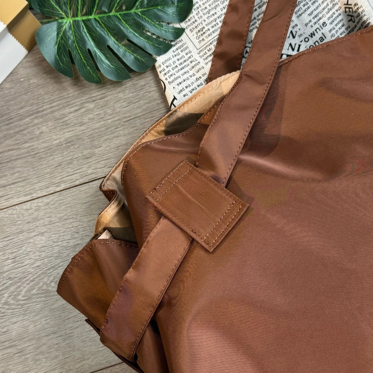 Brown leather bag on a wooden surface with a plant and newspaper in the background